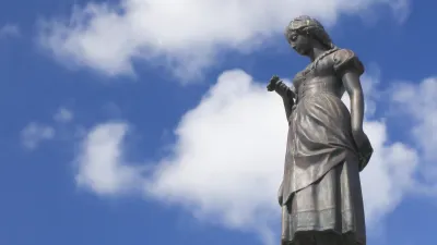 Eine bronzene Statue einer Frau steht auf einem Sockel vor blauem Himmel.