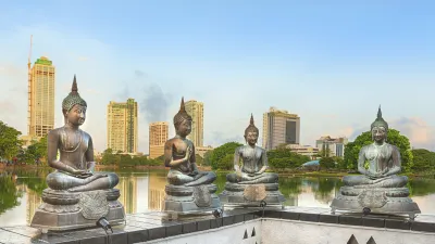 Vier Buddha-Statuen sitzen friedlich vor einem See und modernen Gebäuden.