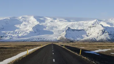 Eine gerade Straße führt durch eine weite Ebene auf schneebedeckte Berge in Island zu.