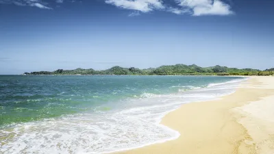 Wellen brechen an einem goldenen Sandstrand mit üppiger Vegetation im Hintergrund.