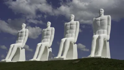 Vier weiße Männerfiguren sitzen auf Sockeln an der Küste.