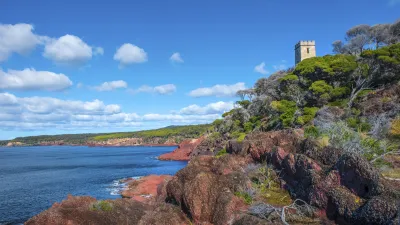 Roter Felsen am Meer mit einem historischen Turm im Hintergrund.