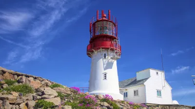 Weißer Leuchtturm mit rotem Dach steht auf felsiger Küste.