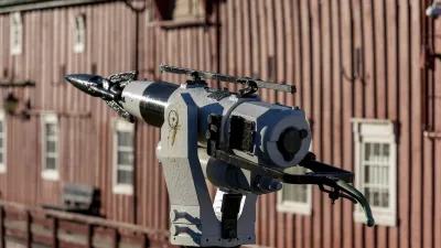 Historische Kamera auf einem Sockel vor einem roten Holzgebäude.