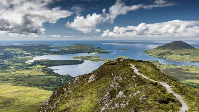 Grüne Hügel und ein verschlungener Pfad führen zu einem Blick auf See und Berge.